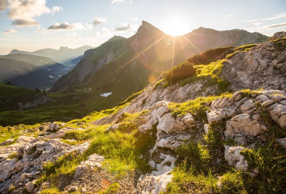 Sonnenaufgang über einer alpinen Berglandschaft mit grünen Wiesen und Felsen.