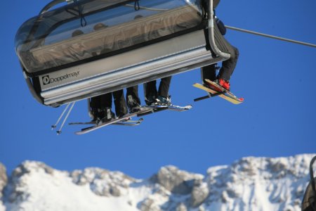 Skifahrer fahren in einem Sessellift, der vor einem schneebedeckten Gebirgshorizont schwebt.