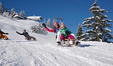 Familie beim Rodeln im Schnee auf einer verschneiten Bergwiese.