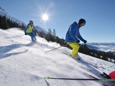 Zwei Skifahrer auf einer Piste in den Alpen bei sonnigem Wetter.