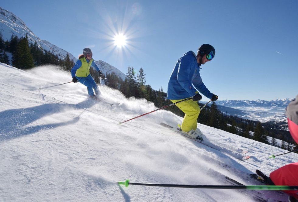 Zwei Skifahrer auf einer Piste in den Alpen bei sonnigem Wetter.