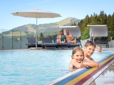 Zwei lachende Kinder am Poolrand mit Bergen und Liegen im Hintergrund.