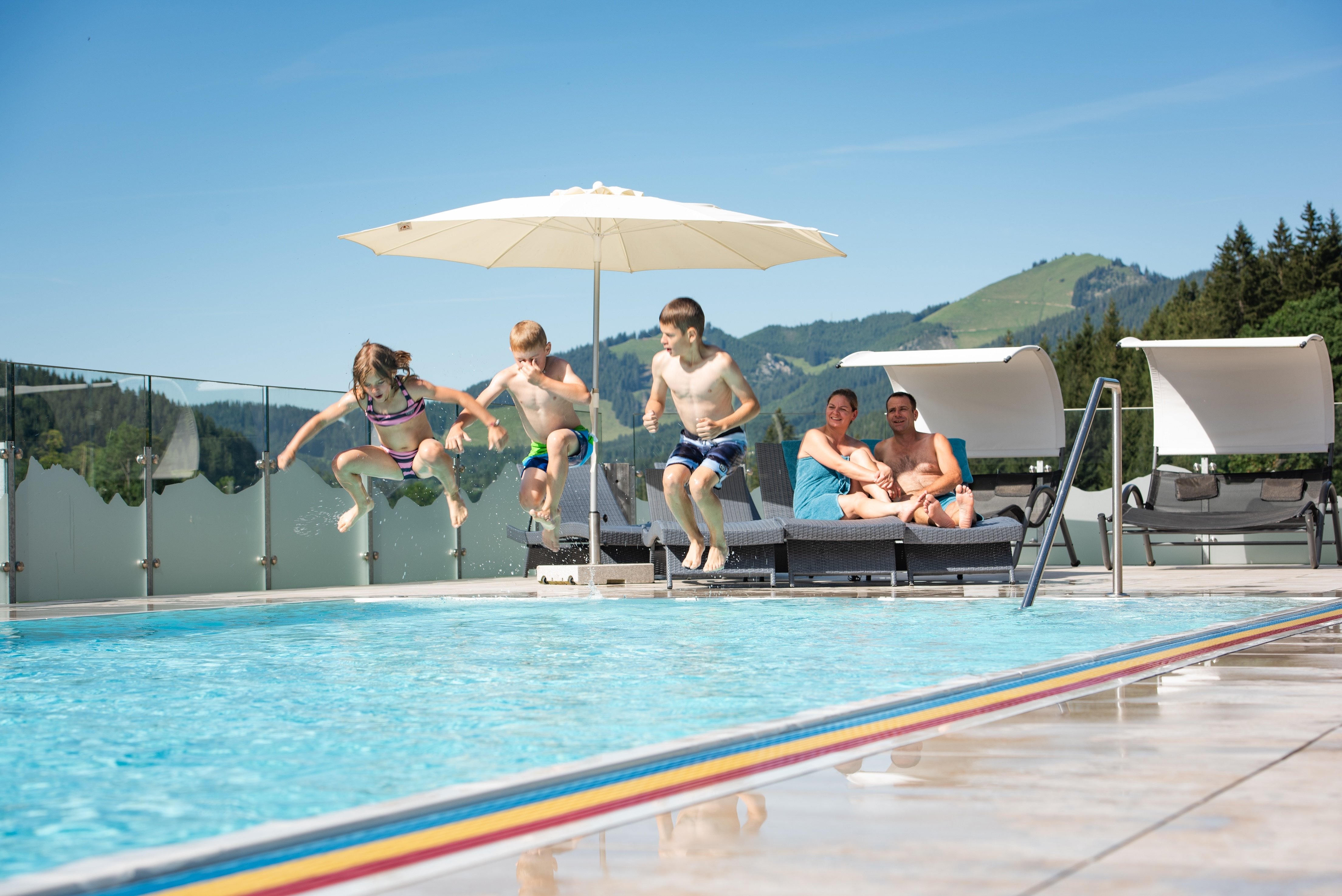 Kinder springen ins Becken vor Bergkulisse und Sonnenliegen.