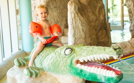 Kleines Kind mit Schwimmflügeln sitzt auf einem Krokodil-Spielzeug im Wasser.