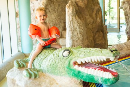 Kleines Kind mit Schwimmflügeln sitzt auf einem Krokodil-Spielzeug im Wasser.