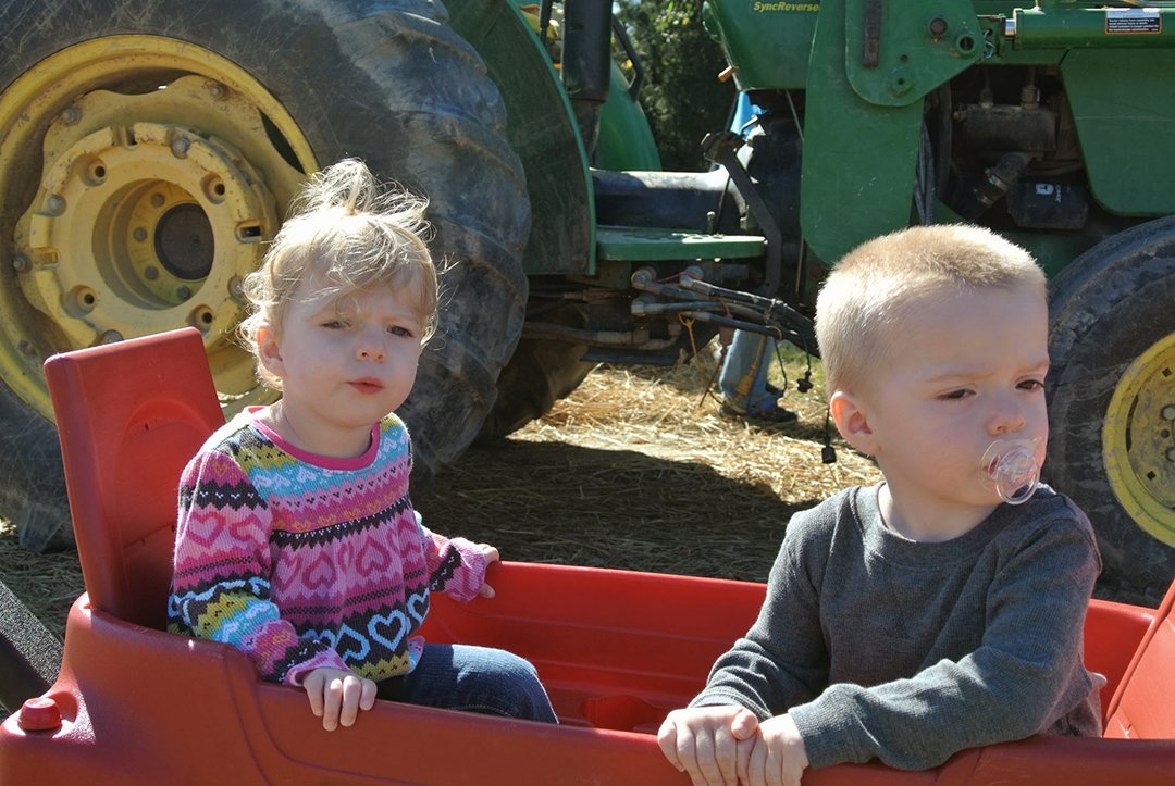 Zwei Kinder sitzen in einem roten Wagen mit einem Traktor im Hintergrund.