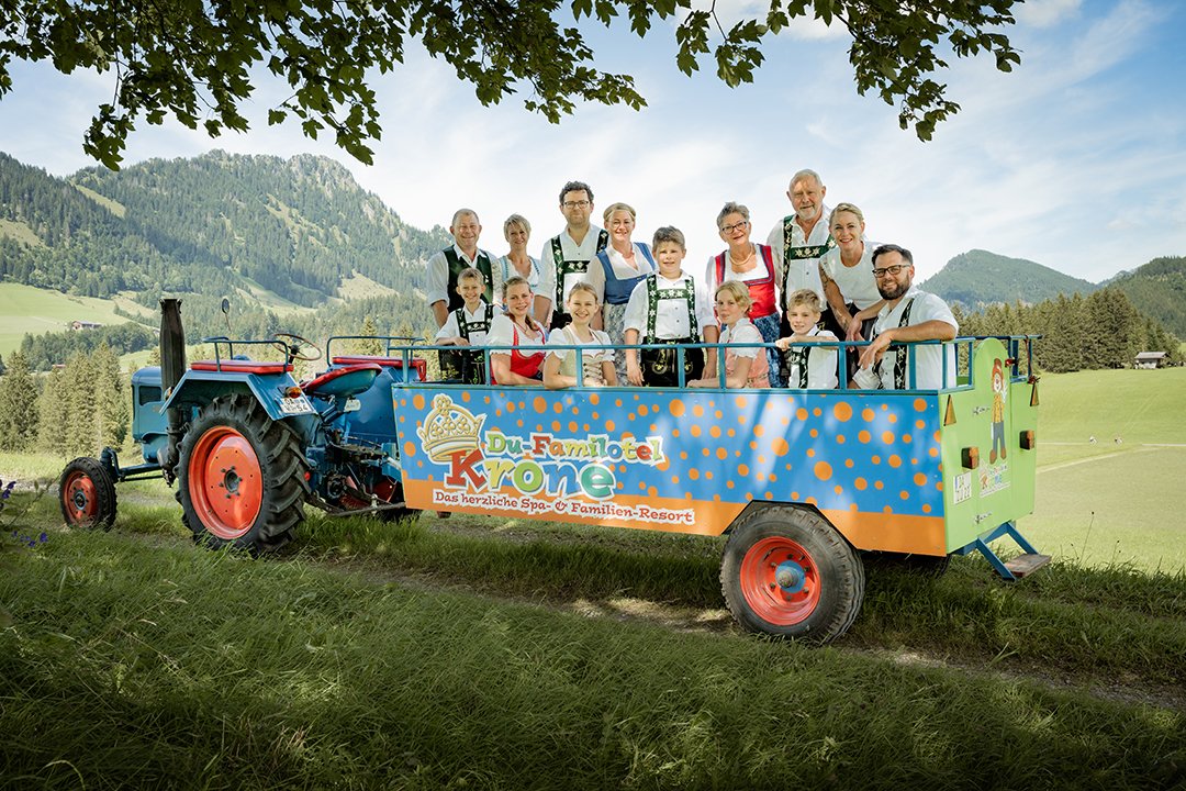 Eine Gruppe von Personen in traditioneller Tracht steht auf einem Traktoranhänger vor einer Berglandschaft.