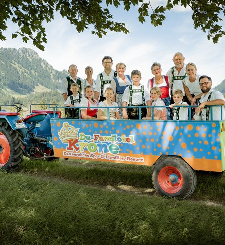 Eine Gruppe von Personen in traditioneller Tracht steht auf einem Traktoranhänger vor einer Berglandschaft.