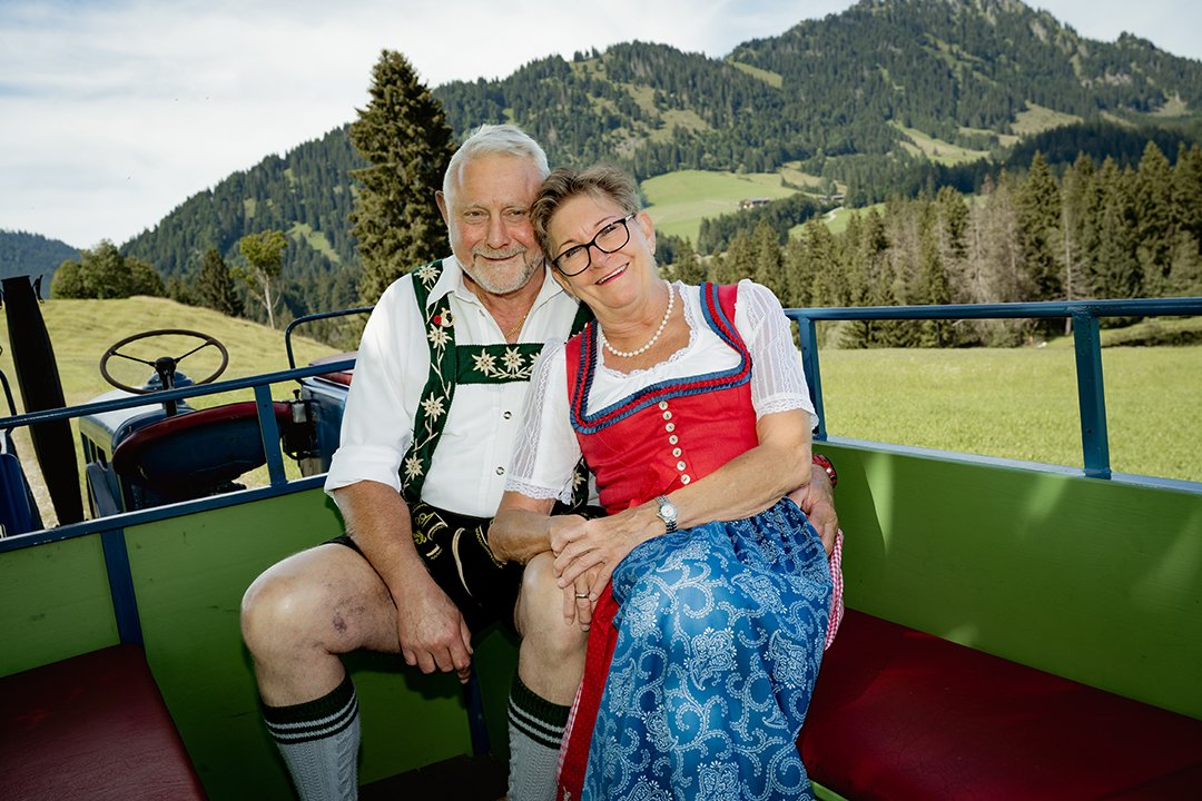 Ein älteres Paar in Trachten sitzt auf einem Traktor vor den Bergen.