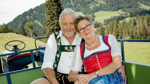 Ein älteres Paar in Trachten sitzt auf einem Traktor vor den Bergen.