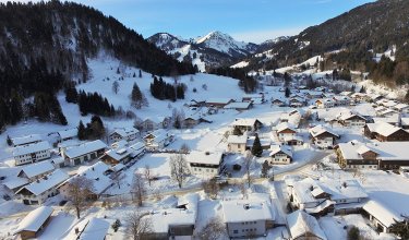 Das Bild zeigt ein verschneites Dorf in den Bergen mit mehreren Häusern und Bäumen.