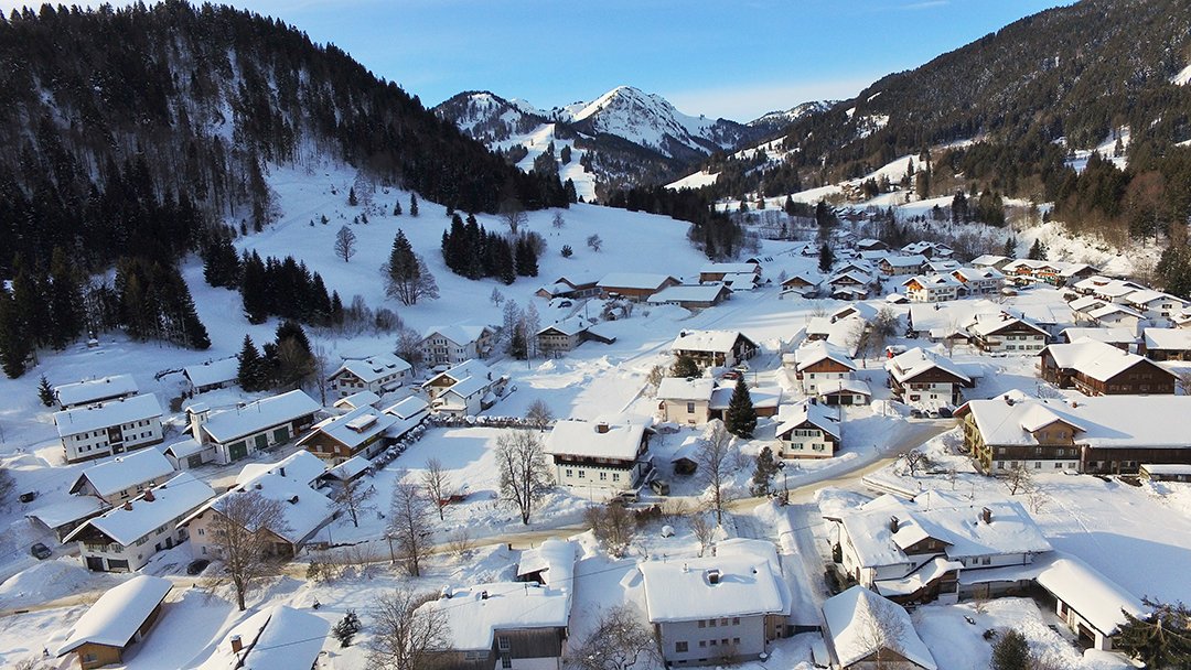 Das Bild zeigt ein verschneites Dorf in den Bergen mit mehreren Häusern und Bäumen.