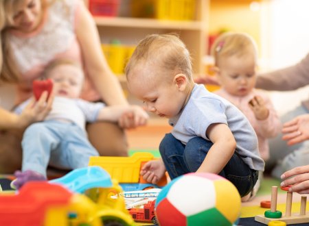 Kleinkind, das mit Spielzeugen auf dem Boden spielt und andere Kinder und Erwachsene im Hintergrund.