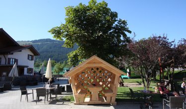 Das Bild zeigt einen Holzpavillon im Freien, umgeben von Bäumen und Sitzgelegenheiten.