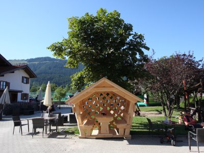 Das Bild zeigt einen Holzpavillon im Freien, umgeben von Bäumen und Sitzgelegenheiten.