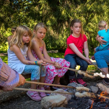 Kinder rösten Stockbrot über einem Lagerfeuer, während Erwachsene dabei helfen.