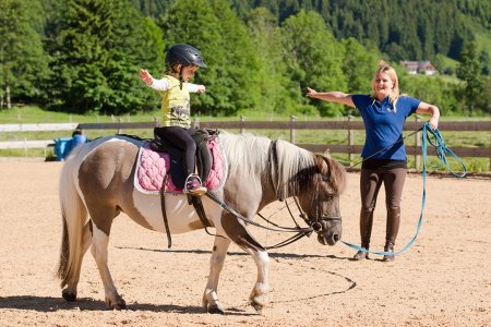 Ein Kind reitet auf einem Pony und streckt beide Arme aus, während es von einer Betreuerin angeleitet wird.
