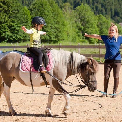 Ein Kind reitet auf einem Pony und streckt beide Arme aus, während es von einer Betreuerin angeleitet wird.