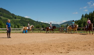 Reiterinnen reiten in einer Reitbahn, während eine Instruktorin auf dem Boden steht.