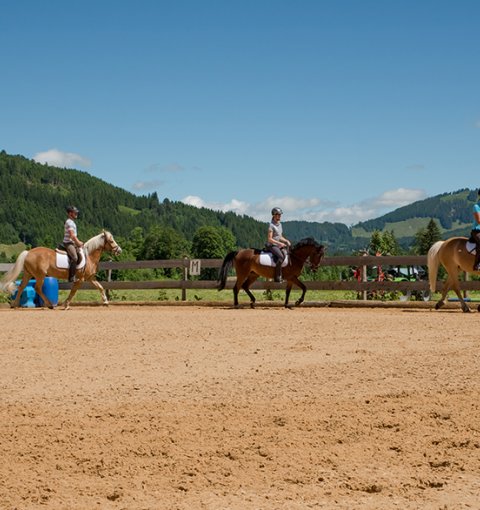 Reiterinnen reiten in einer Reitbahn, während eine Instruktorin auf dem Boden steht.