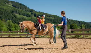 Zwei Kinder reiten zusammen auf einem Pony, das von einer Betreuerin geführt wird.