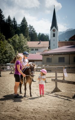 Kind auf Pony wird geführt, weiteres Kind schaut zu.