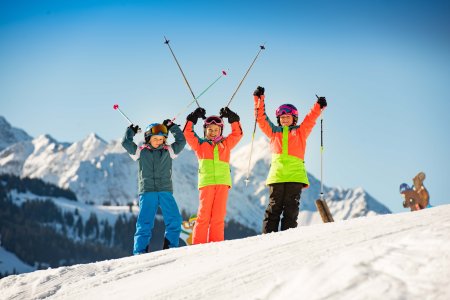 Drei Mädchen mit bunten Skianzügen auf Skiern in der Schneelandschaft halten Skistöcke in die Höhe.