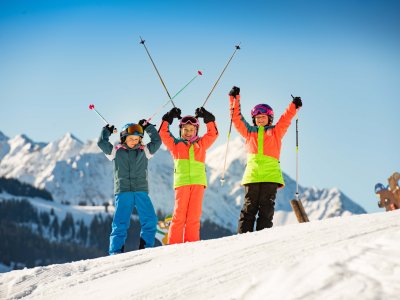 Drei Mädchen mit bunten Skianzügen auf Skiern in der Schneelandschaft halten Skistöcke in die Höhe.