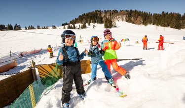 Drei Kinder in Skimontur auf der Skipiste zeigen Daumen hoch in die Kamera.