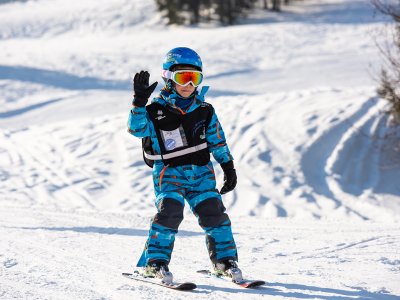 Kind in blauem Anzug fährt Ski und winkt auf sonniger Piste