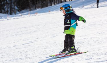Kind fährt alleine Ski auf sonniger Piste mit Wald im Hintergrund