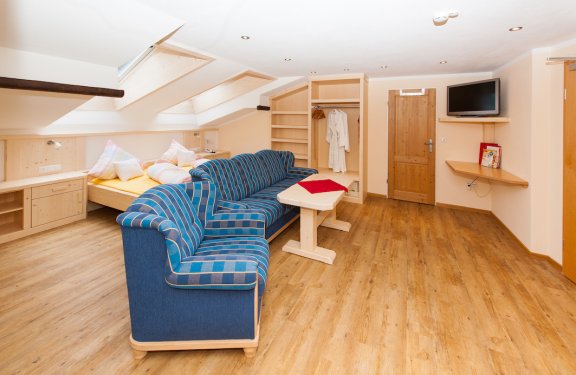 Großes Doppelzimmer mit blauem Sofa, Couchtisch, Kasten und Fernseher.
