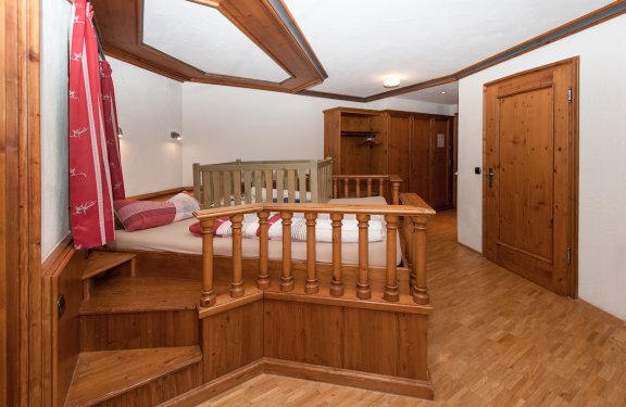 Seitenansicht von Doppelbett auf einem Holzpodest mit Holzpfeilern rundherum und Babybettaufsatz darauf.