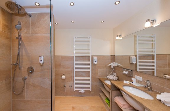 Badezimmer mit beigen Fliesen, zwei Waschbecken, ebenerdiger Dusche und Handtuchwärmer.