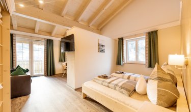 Doppelbett wird durch Wand zu Wohnbereich mit Sofa, Sitzecke und Fernseher getrennt.