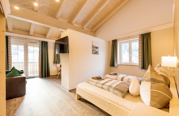 Doppelbett wird durch Wand zu Wohnbereich mit Sofa, Sitzecke und Fernseher getrennt.